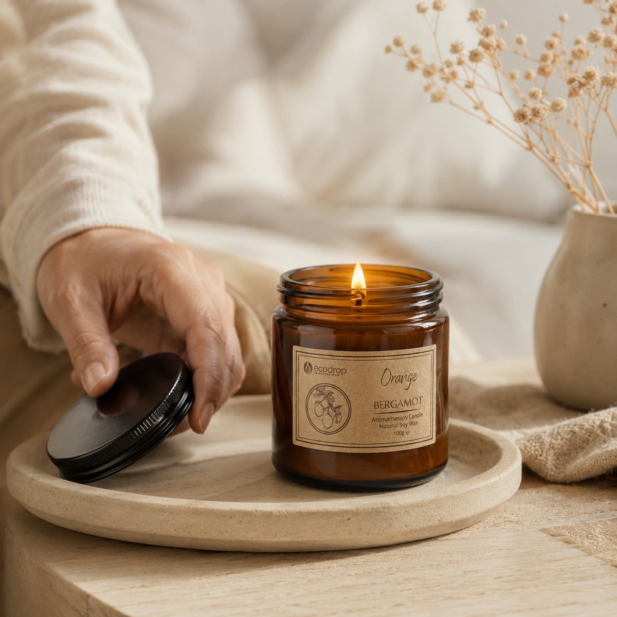Hand holding the black lid beside a lit Orange & Bergamot candle on a round stone tray